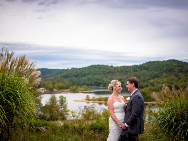 A bride and groom stand in a scenic outdoor setting with a lake and hills in the background, framed by tall grasses.