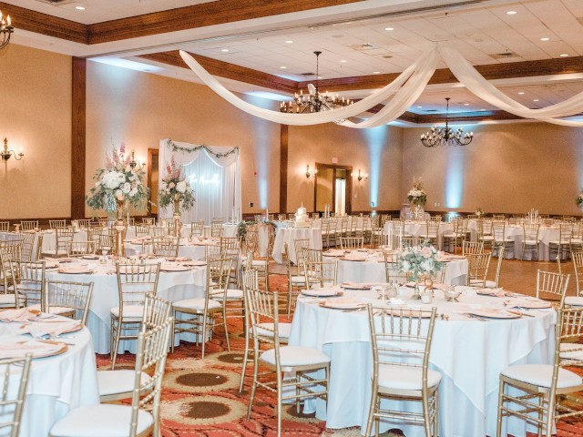 A decorated banquet hall features round tables set with white linens and gold chairs, elegant chandeliers, and draped fabric on the ceiling.