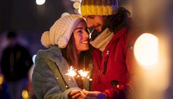 A couple in winter attire holds sparklers, standing close together amidst blurry festive lights, smiling warmly at each other.