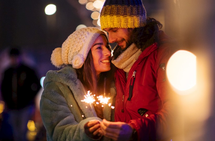 A couple in winter attire holds sparklers, standing close together amidst blurry festive lights, smiling warmly at each other.