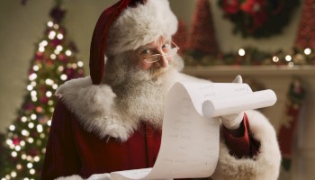 Santa Claus is holding a long scroll, checking his list, with a decorated Christmas tree and lights in the background.