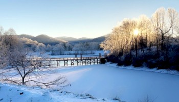 A snow-covered landscape with a frozen lake, bare trees, and a wooden bridge, under a clear sky with sunlight streaming through the trees.