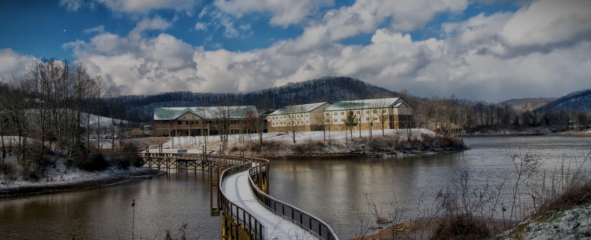 A scenic view of a lake with ice, a curved walkway, and buildings on the shoreline, surrounded by snowy hills under a cloudy sky.