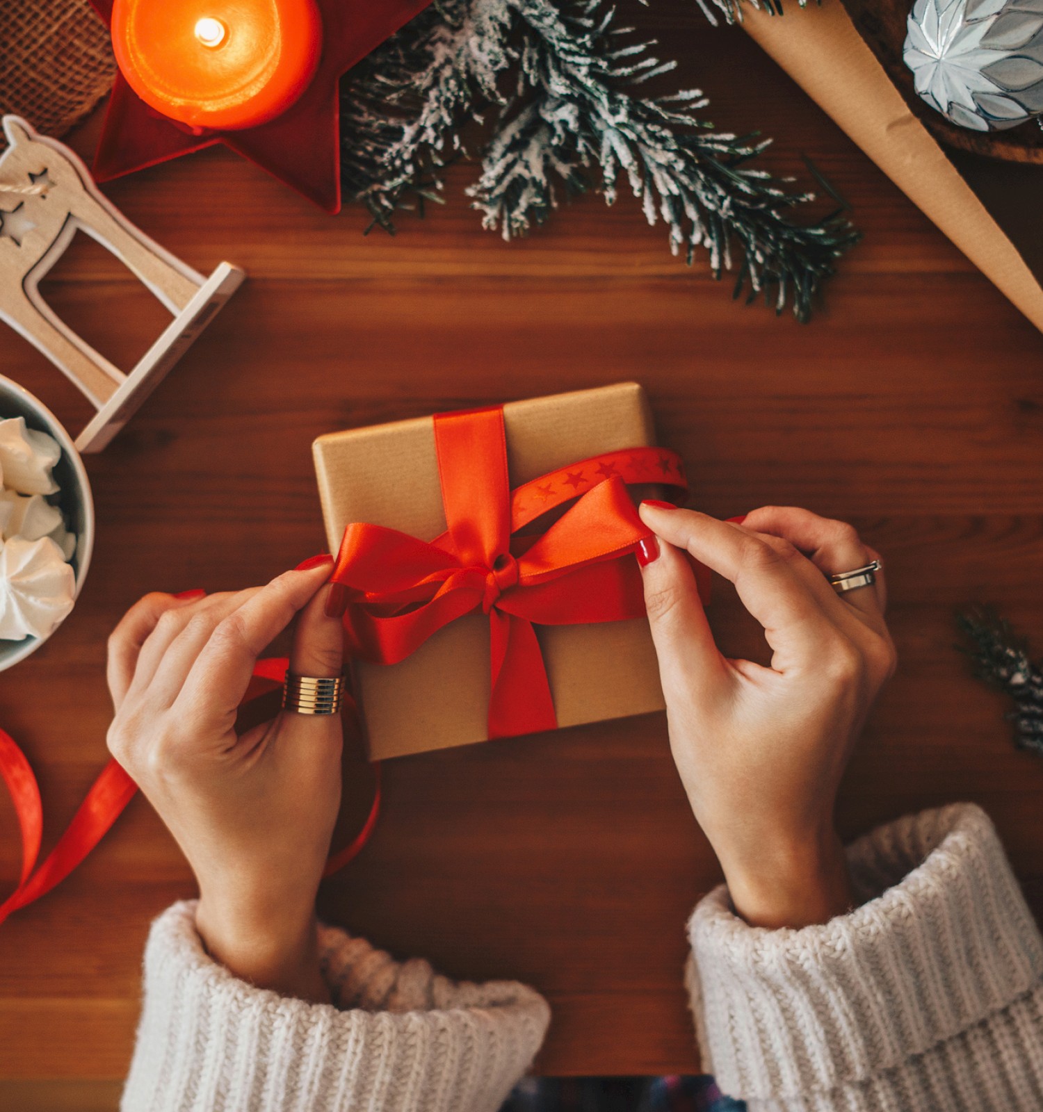 The image shows someone wrapping a gift with a red bow, surrounded by festive items like candles, marshmallows, and holiday decorations.