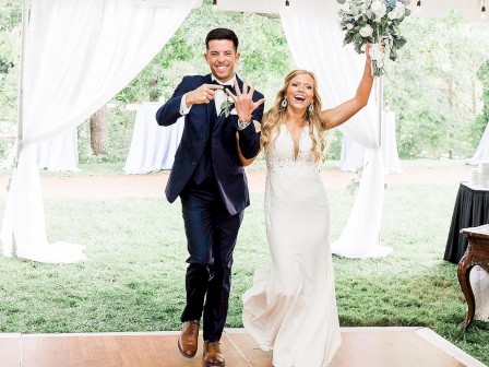 A couple, dressed formally, joyfully enters a tented area; the woman holds a bouquet, the man gestures enthusiastically.