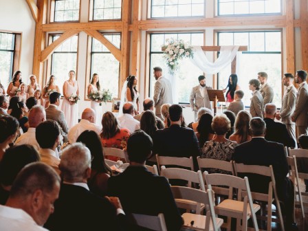 A wedding ceremony inside a wooden venue with large windows, people seated, and a bridal party at the front.