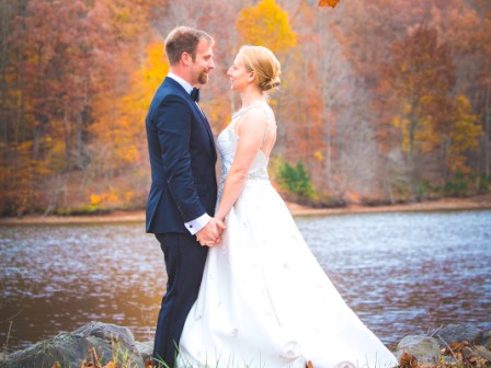 A couple in wedding attire stands by a lake, surrounded by autumn foliage, holding hands and gazing at each other.