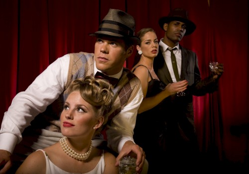 Four people dressed in vintage attire with hats and dresses pose against a red curtain backdrop, holding drinks.