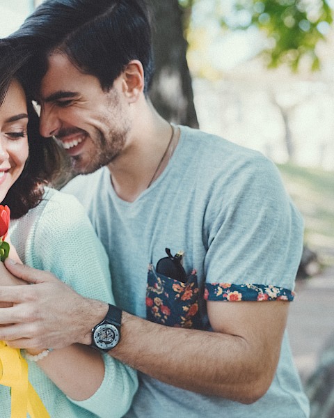 A couple is in a park; the man hugs the woman from behind as she holds red flowers, both smiling warmly.