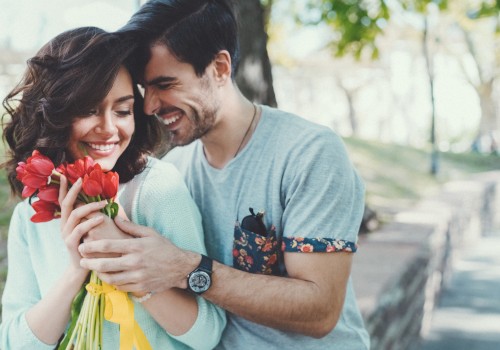 A couple is in a park; the man hugs the woman from behind as she holds red flowers, both smiling warmly.