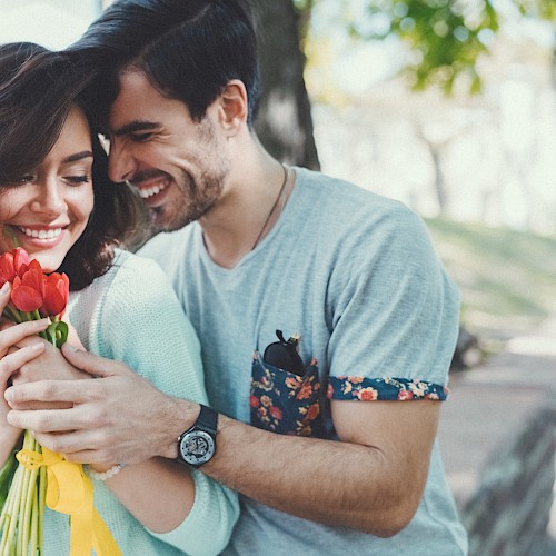 A couple is in a park; the man hugs the woman from behind as she holds red flowers, both smiling warmly.