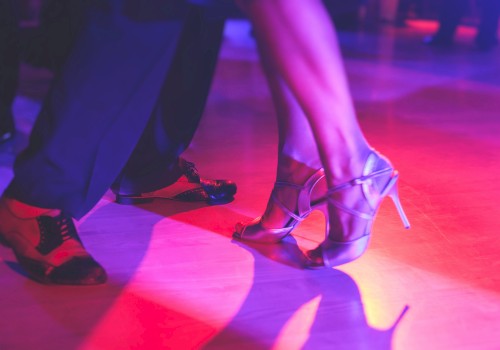 Two pairs of feet are dancing on a colorful, illuminated floor—one in heels and the other in dress shoes.