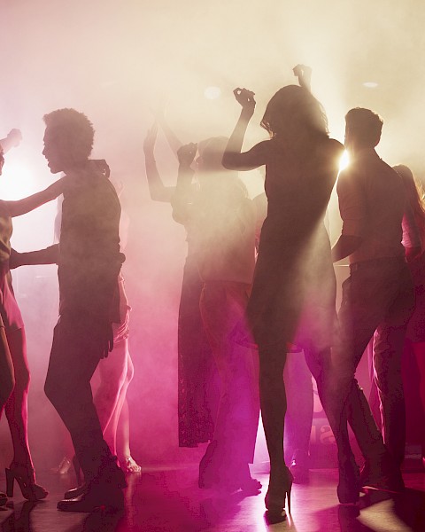 A group of people dancing in a dimly lit, foggy environment with colorful lights and silhouettes visible.