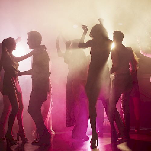 A group of people dancing in a dimly lit, foggy environment with colorful lights and silhouettes visible.