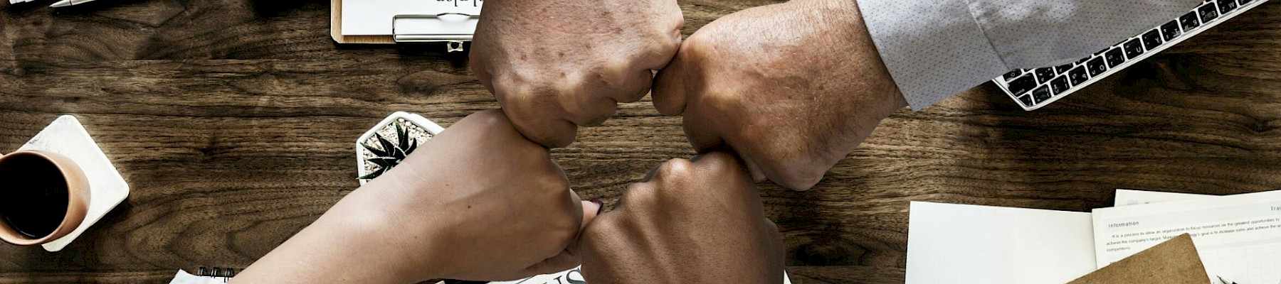 Four hands fist-bumping over a desk with business documents, a calculator, a laptop, and a phone, symbolizing teamwork.