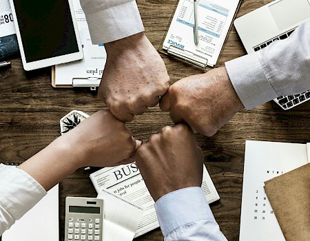 Four hands fist-bumping over a desk with business documents, a calculator, a laptop, and a phone, symbolizing teamwork.