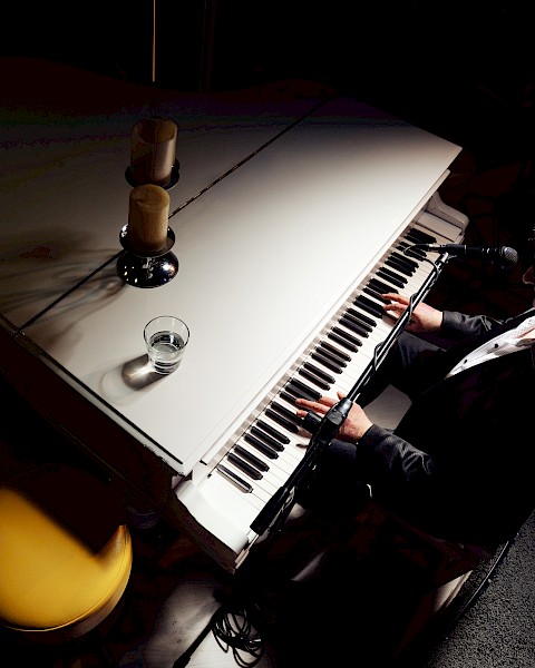 A person is playing a white grand piano, with a glass and two candles on top, in a dimly lit setting.