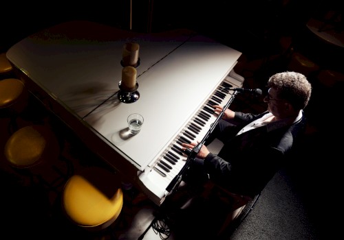 A person is playing a white grand piano, with a glass and two candles on top, in a dimly lit setting.