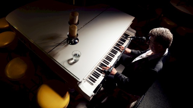 A person is playing a white grand piano, with a glass and two candles on top, in a dimly lit setting.