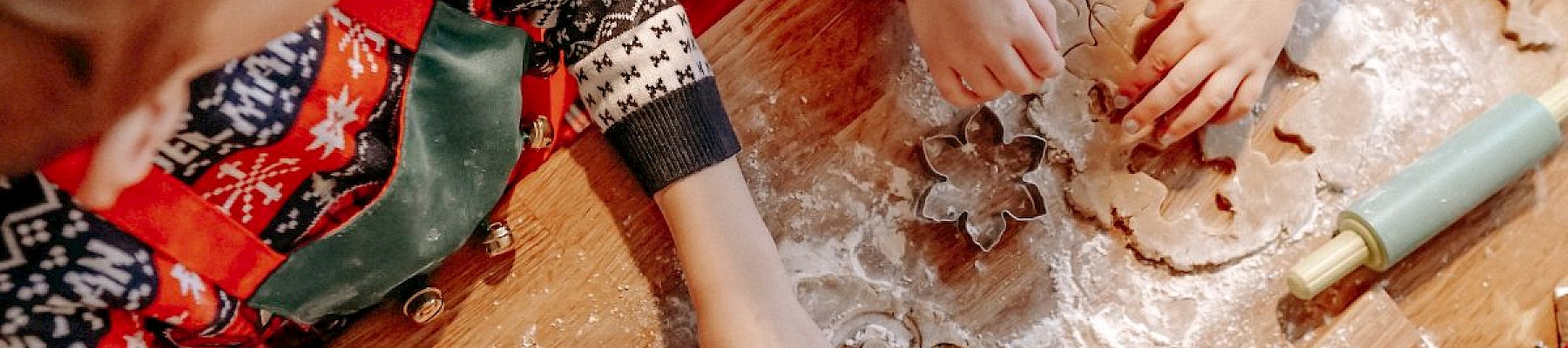 Two children in festive clothes are using cookie cutters on dough on a floured table, with a rolling pin nearby.