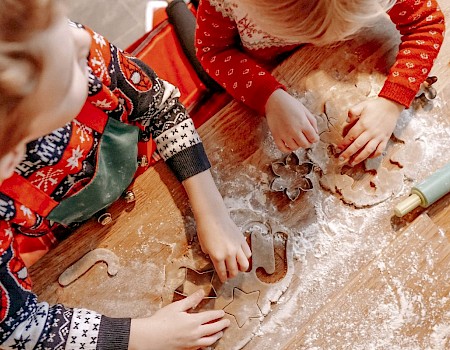 Two children in festive clothes are using cookie cutters on dough on a floured table, with a rolling pin nearby.