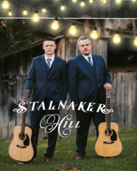 Two people in suits stand with guitars in front of a rustic building, with 