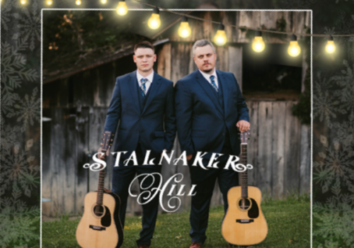 Two people in suits stand with guitars in front of a rustic building, with 