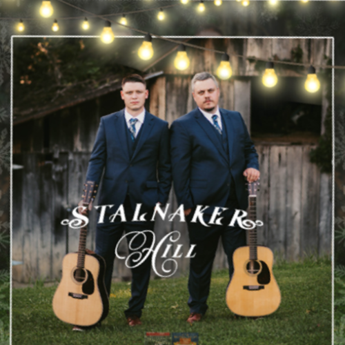 Two people in suits stand with guitars in front of a rustic building, with 