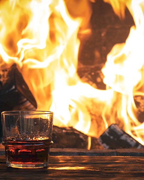 A glass of whiskey and a bottle stopper sit on a table in front of a roaring fireplace, creating a cozy ambiance.