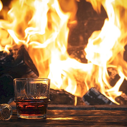 A glass of whiskey and a bottle stopper sit on a table in front of a roaring fireplace, creating a cozy ambiance.