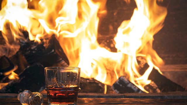 A glass of whiskey and a bottle stopper sit on a table in front of a roaring fireplace, creating a cozy ambiance.