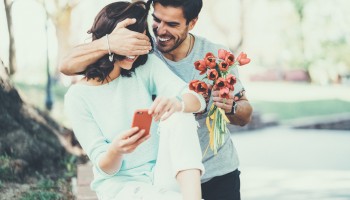 A person surprises another by covering their eyes while holding a bouquet of flowers. The surprised person holds a phone and smiles.