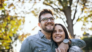 A couple is smiling and embracing in an outdoor setting with trees and sunlight in the background.