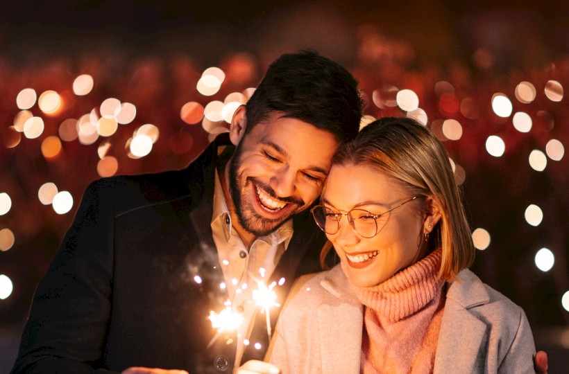 A couple is joyfully holding sparklers at night, surrounded by blurry festive lights, creating a romantic ambiance.