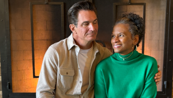 A couple sits together near a fireplace. The man wears a beige shirt, and the woman is in a green sweater, looking at each other warmly.