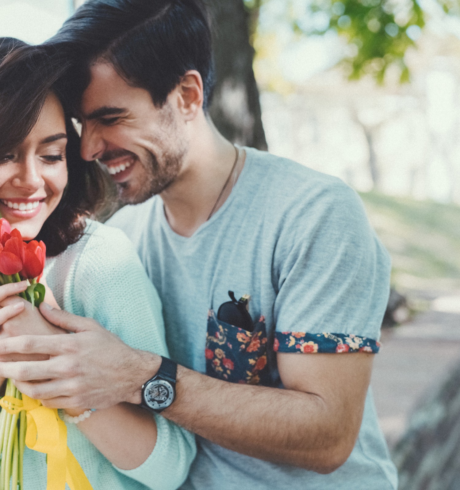 A couple is smiling and embracing outdoors, with the woman holding red flowers. They appear happy and affectionate in a natural setting.