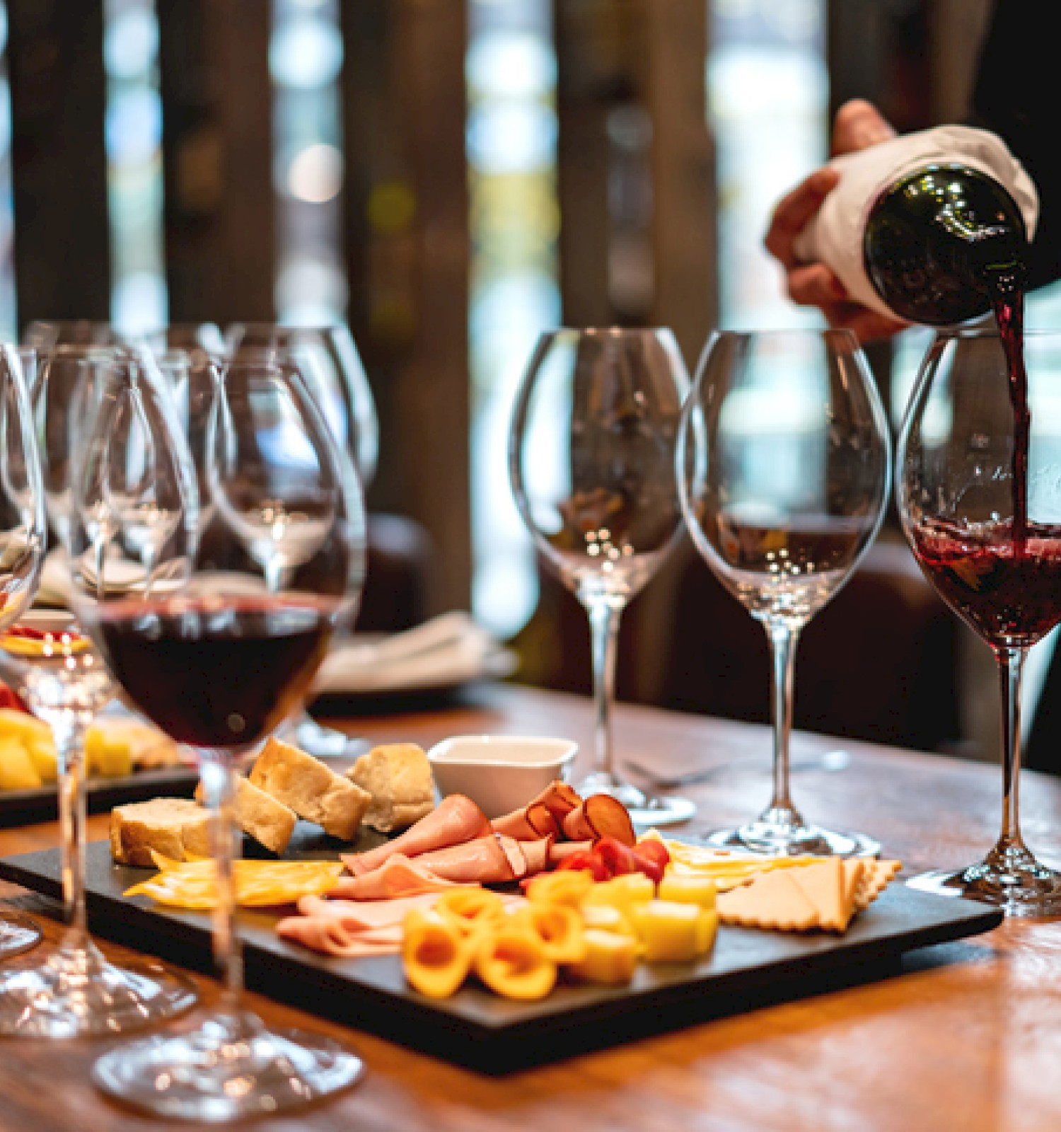 The image shows a wine tasting setup with glasses of red wine and platters of cheese and meats on a wooden table.