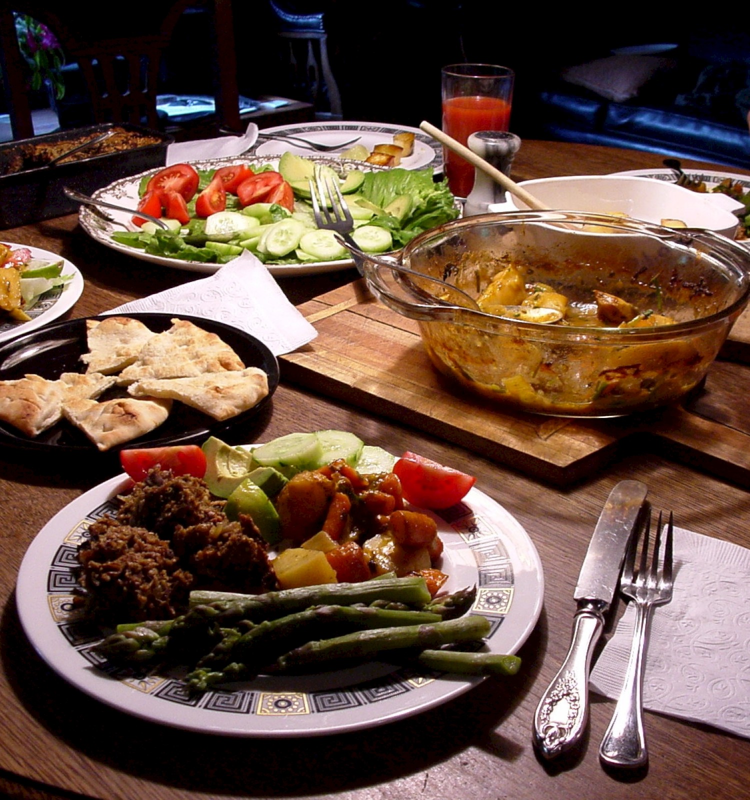 A dining table with a variety of foods including salad, bread, a cooked dish, asparagus, and drinks, set for a meal.