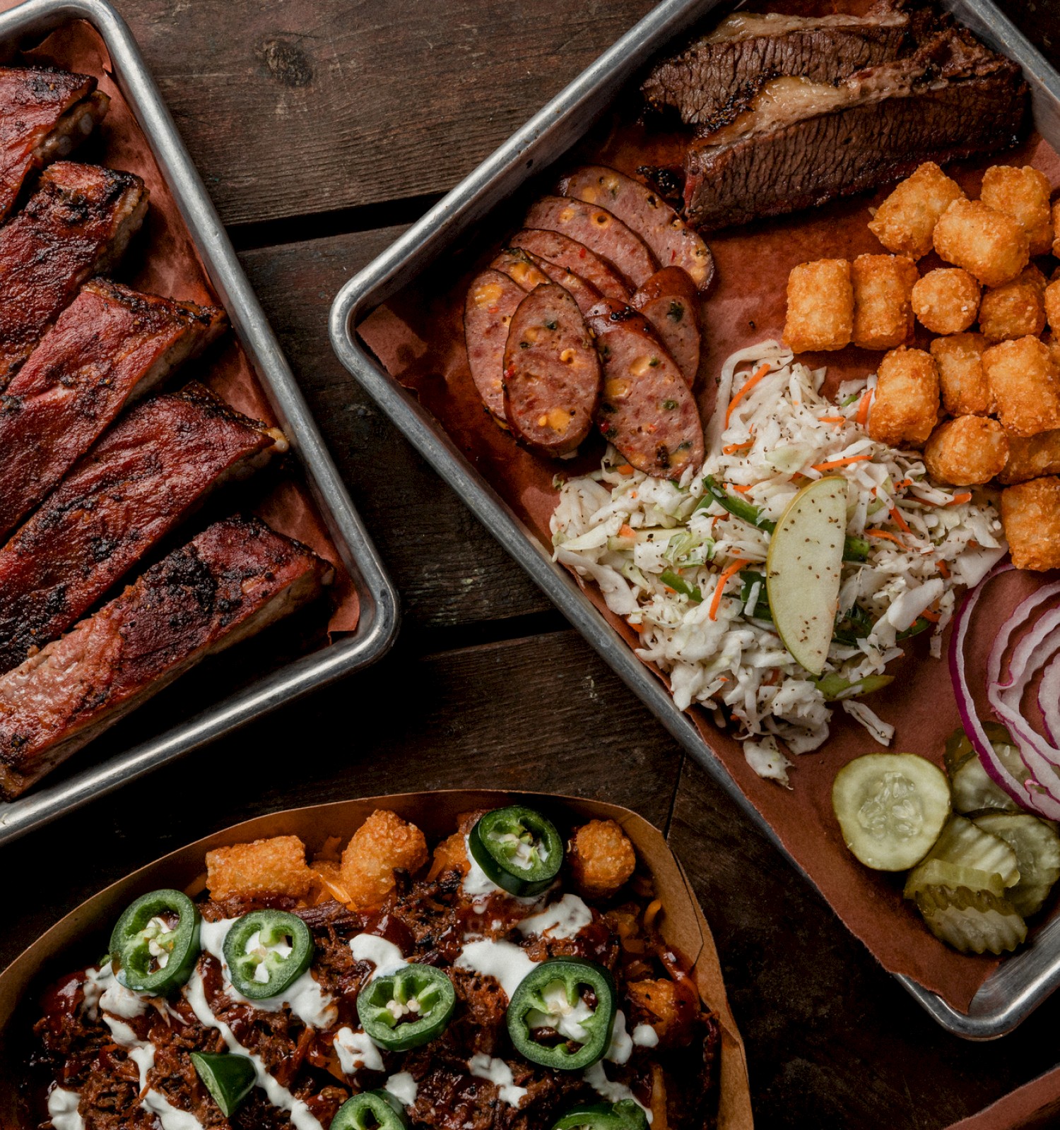 Assorted barbecue dishes with ribs, sausages, coleslaw, tater tots, nachos, jalapeños, pickles, and onions on trays.