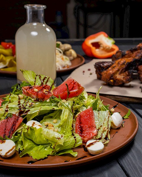 A fresh salad with leafy greens, grapefruit, mozzarella, and balsamic drizzle, alongside roasted meat and a bottle of lemonade.