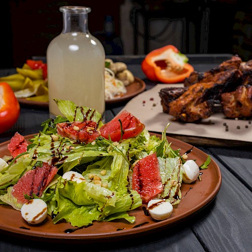 A fresh salad with leafy greens, grapefruit, mozzarella, and balsamic drizzle, alongside roasted meat and a bottle of lemonade.