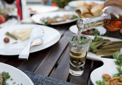 A person pours a clear liquid from a bottle into a glass on a table with plates of food, including pickles and cheese.