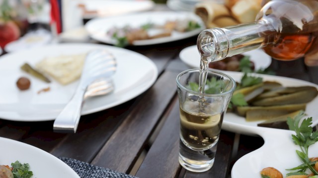 A person pours a clear liquid from a bottle into a glass on a table with plates of food, including pickles and cheese.