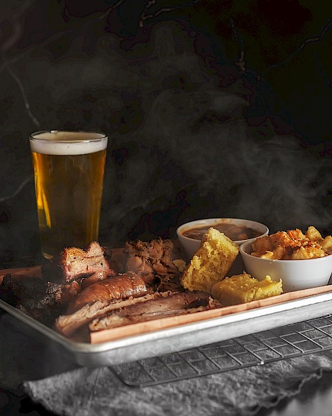 The image shows a tray of smoked meats, various sides, and a glass of beer, creating a hearty meal setting.