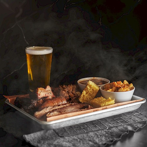 The image shows a tray of smoked meats, various sides, and a glass of beer, creating a hearty meal setting.