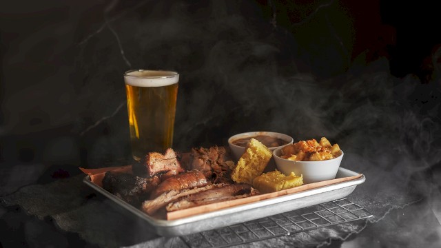 The image shows a tray of smoked meats, various sides, and a glass of beer, creating a hearty meal setting.