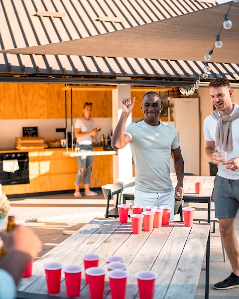 People are playing beer pong outdoors, enjoying a sunny day. There are red cups on the table and a modern outdoor kitchen behind them.