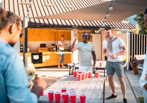 People are playing beer pong outdoors, enjoying a sunny day. There are red cups on the table and a modern outdoor kitchen behind them.