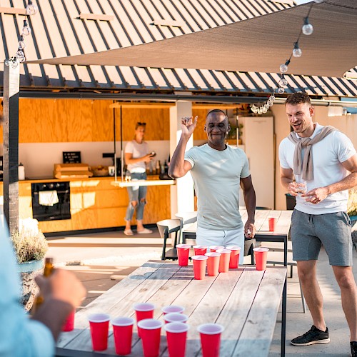 People are playing beer pong outdoors, enjoying a sunny day. There are red cups on the table and a modern outdoor kitchen behind them.