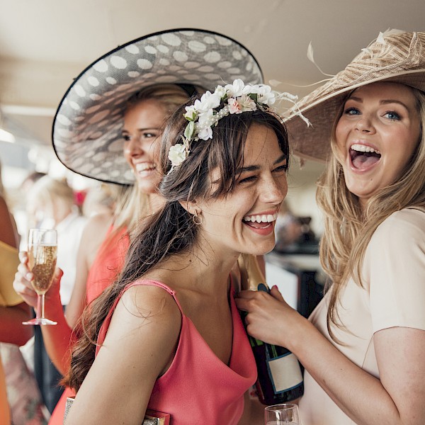 A group of women in stylish hats and dresses enjoy a joyful moment together, while holding glasses of champagne at a social event.